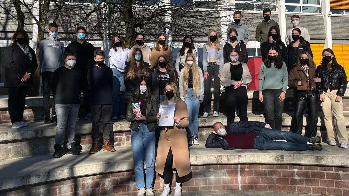 Die Klasse 9b mit Schulleiterin Birgitta Pieters (links) und Mathe-Lehrer Dr. Michael Wagener, der sich auf seiner Klasse dazu gelegt hat. Im Vordergrund halten zwei Schülerinnen der Klasse die Urkunde und den Buddy-Bär-Pokal in die Höhe.